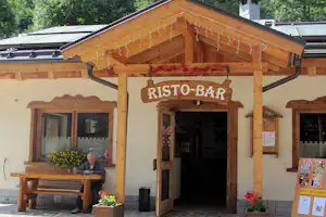 Eingang zur Risto-Bar am Campingplatz mit Holzüberdachung, Sitzbank, Tisch und Blumen vor dem Gebäude.