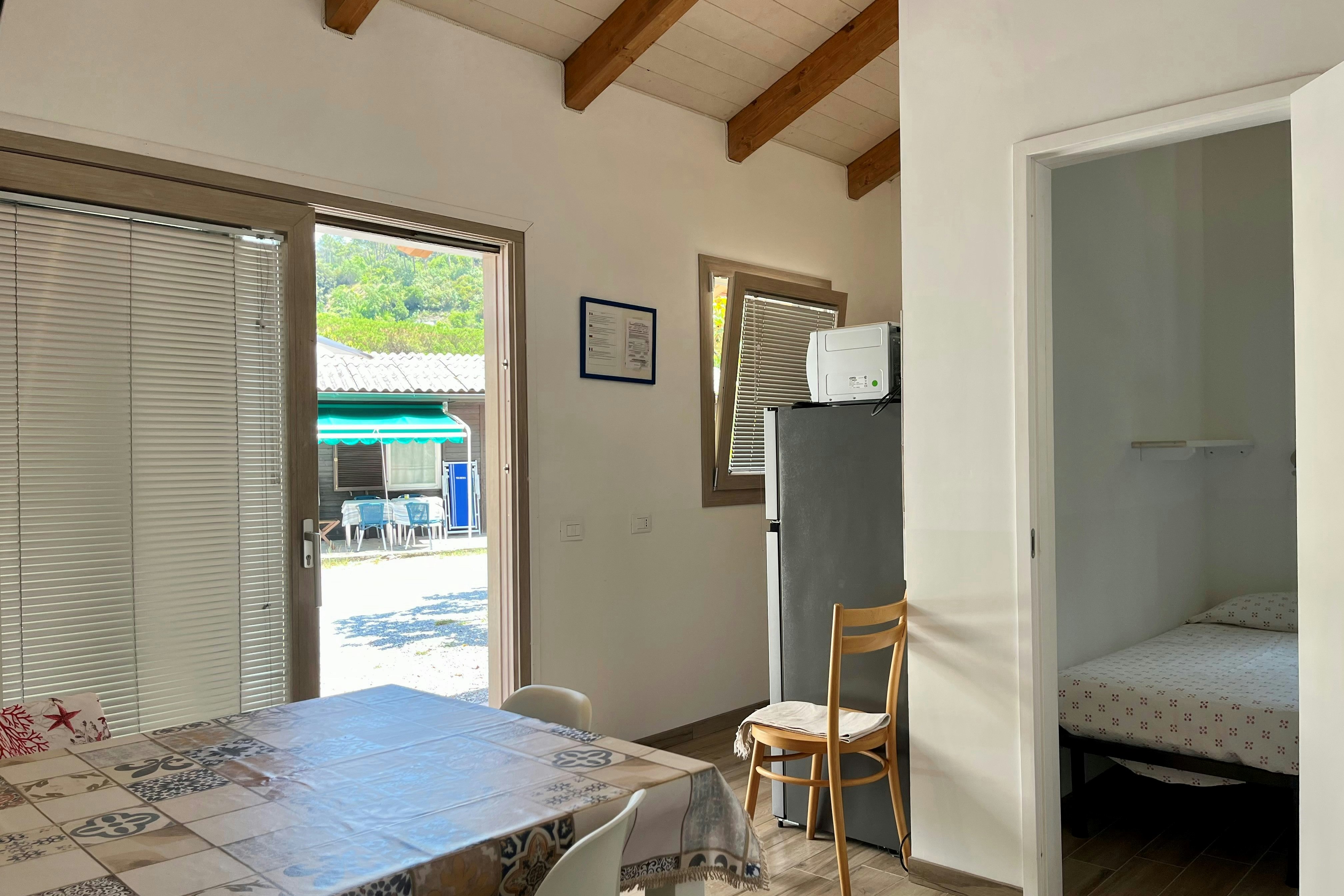 Innenansicht eines Mobilheims mit Bett, Stuhl und Tisch; offene Terrassentür zeigt den Außenbereich mit Sonnenschirm.