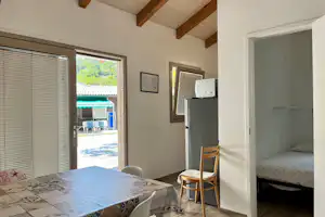 Innenansicht eines Mobilheims mit Bett, Stuhl und Tisch; offene Terrassentür zeigt den Außenbereich mit Sonnenschirm.
