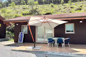 Bungalow mit Terrasse, Sonnenschirm und Tischgruppe vor dem Eingang, Kiesweg und Bäume im Hintergrund.