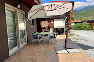 Terrasse am Campingplatz mit Tisch und Stühlen unter einem Sonnenschirm, neben dem Wohnwagen und am Weg.
