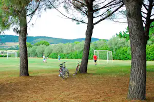 Fußballfeld im Campingbereich mit Toren, Fahrrädern am Boden und Bäumen im Vordergrund, dahinter grüne Wiesen und Hügel.