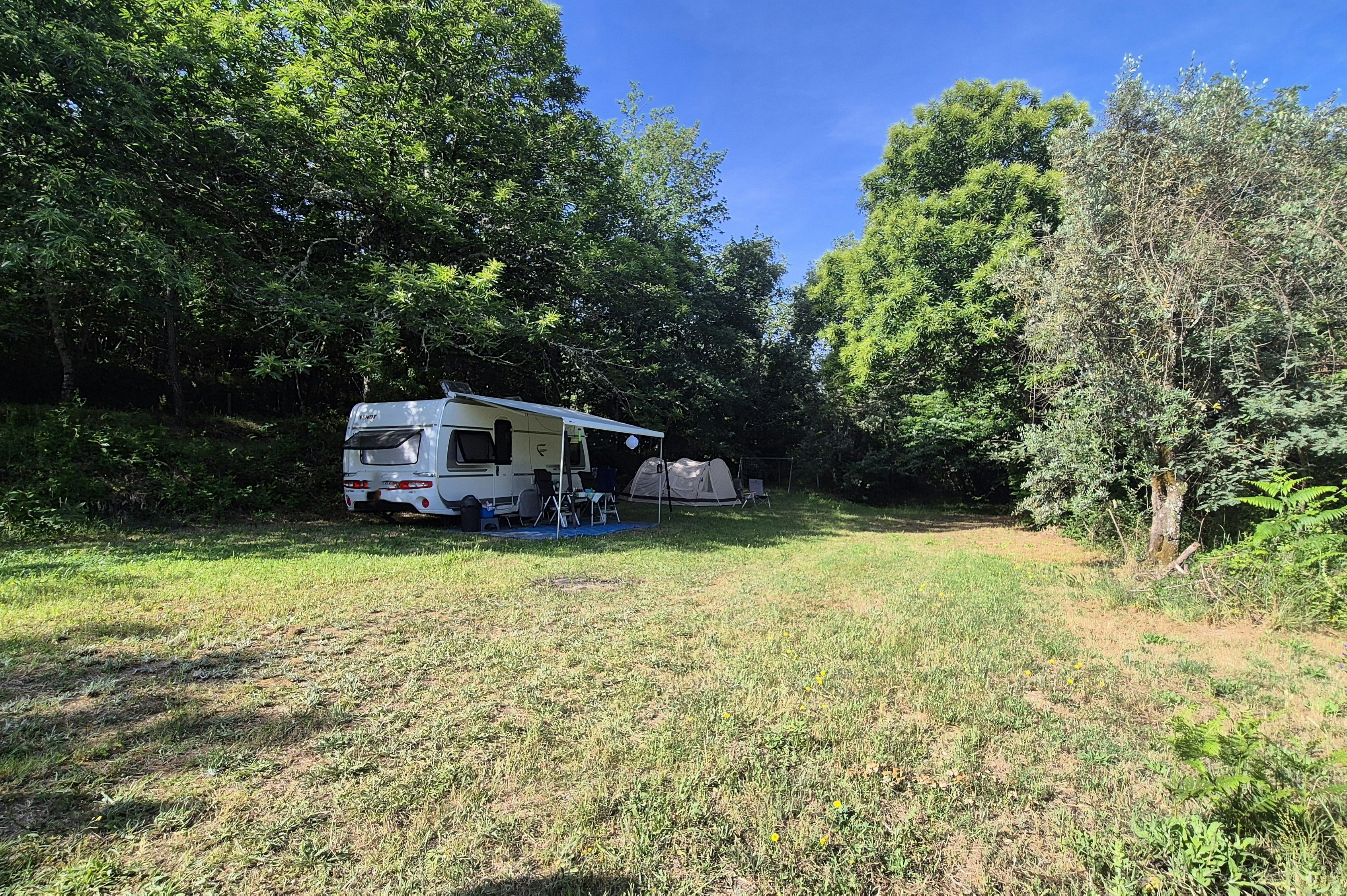 Wohnwagen mit Terrasse und Campingstühlen auf einer Wiese, umgeben von Bäumen im Grünen.