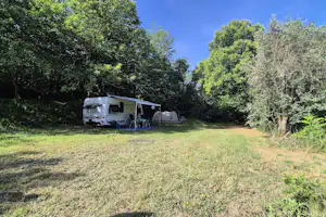 Wohnwagen mit Terrasse und Campingstühlen auf einer Wiese, umgeben von Bäumen im Grünen.