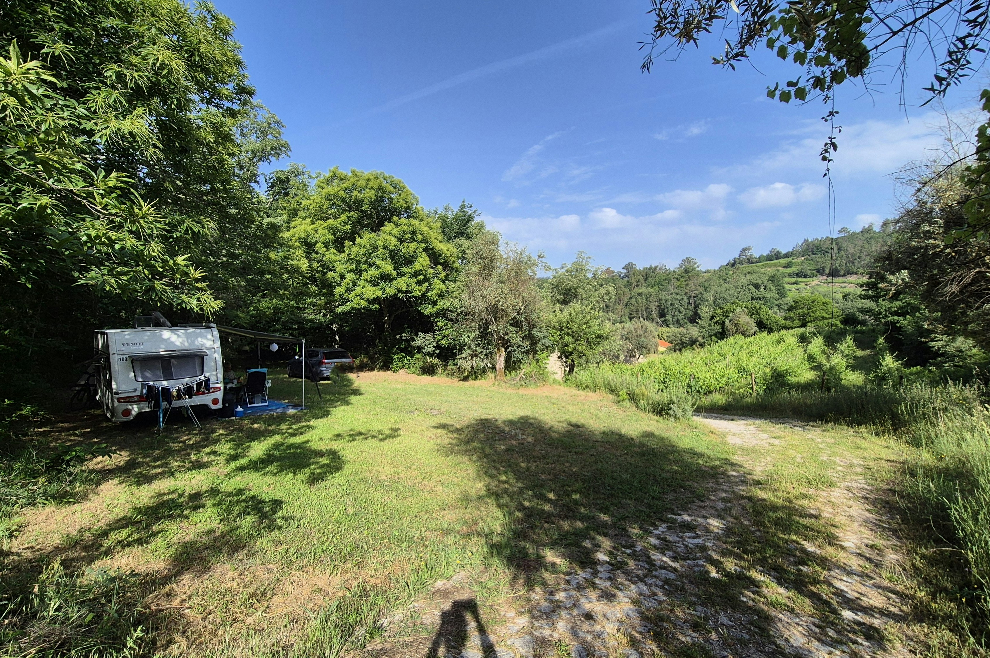 Rasenfläche auf einem Campingplatz mit Wohnwagen und Anhänger, umgeben von Bäumen und grünen Hügeln im Hintergrund.