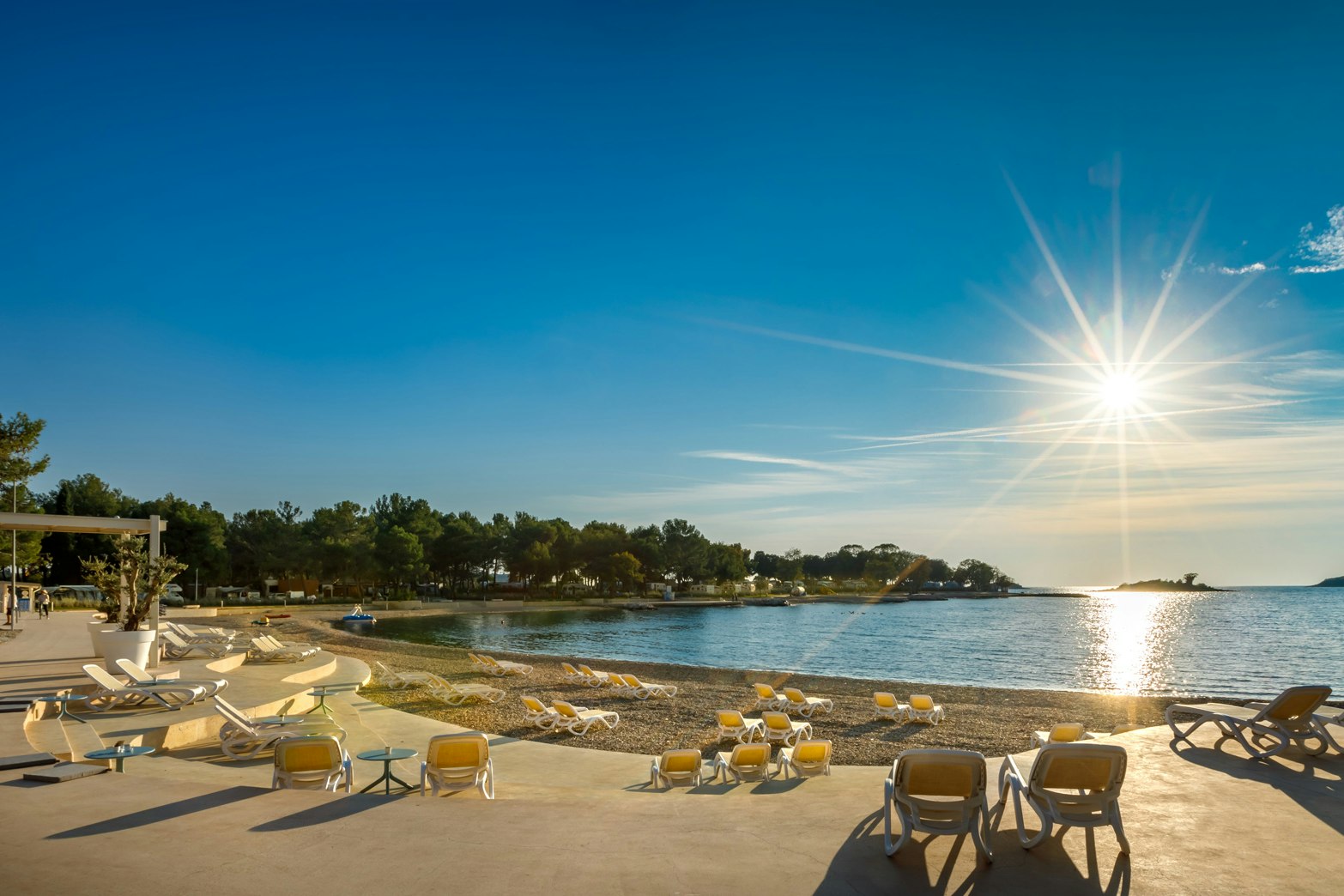 Sonnenuntergang am Strand: Liegen und Sonnenschirme auf einer Terrasse am Ufer, Wasser und Küste im Hintergrund.