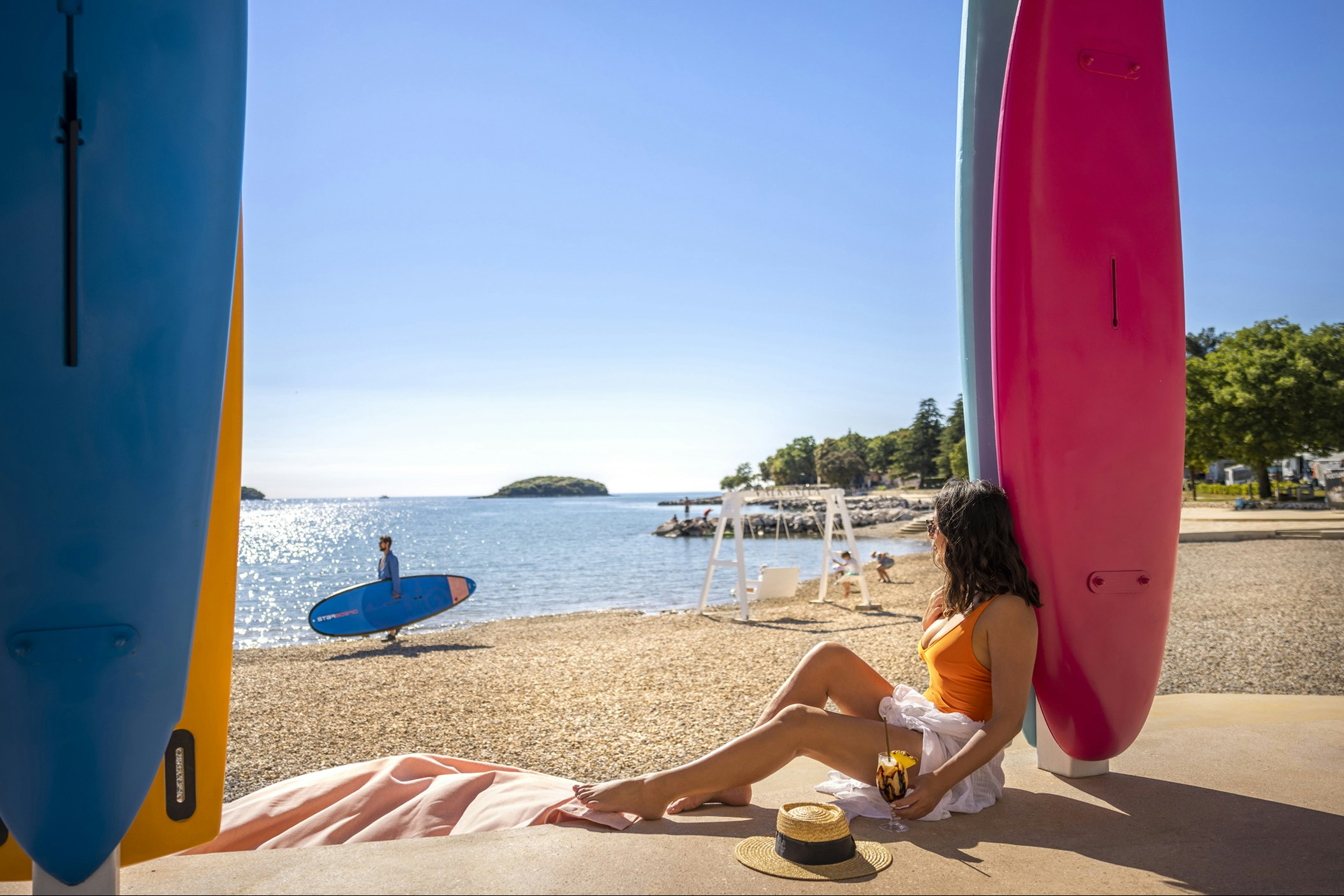 Campingplatz am Meer: Zwei Surfboards am Stellplatz, daneben sitzt eine Person auf einer Liege am Strandweg.