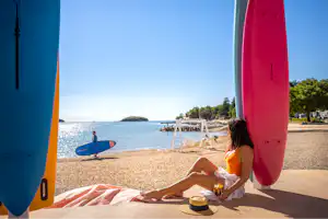 Campingplatz am Meer: Zwei Surfboards am Stellplatz, daneben sitzt eine Person auf einer Liege am Strandweg.