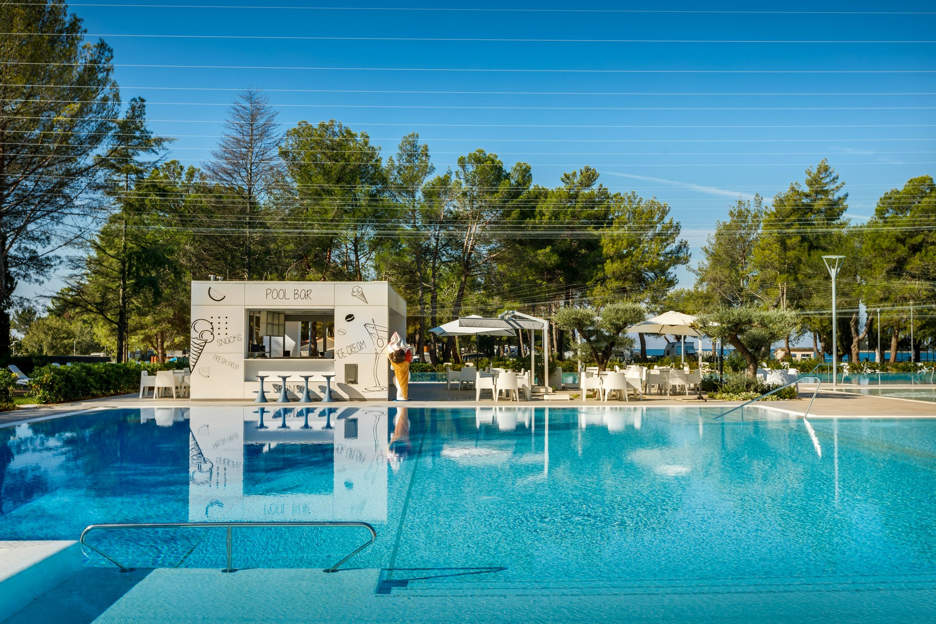 Schwimmbecken mit Liegen und Sonnenschirmen, im Hintergrund ein Gebäude und Bäume auf dem Campingplatz.