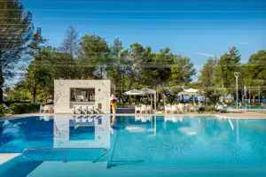 Schwimmbecken mit Liegen und Sonnenschirmen, im Hintergrund ein Gebäude und Bäume auf dem Campingplatz.
