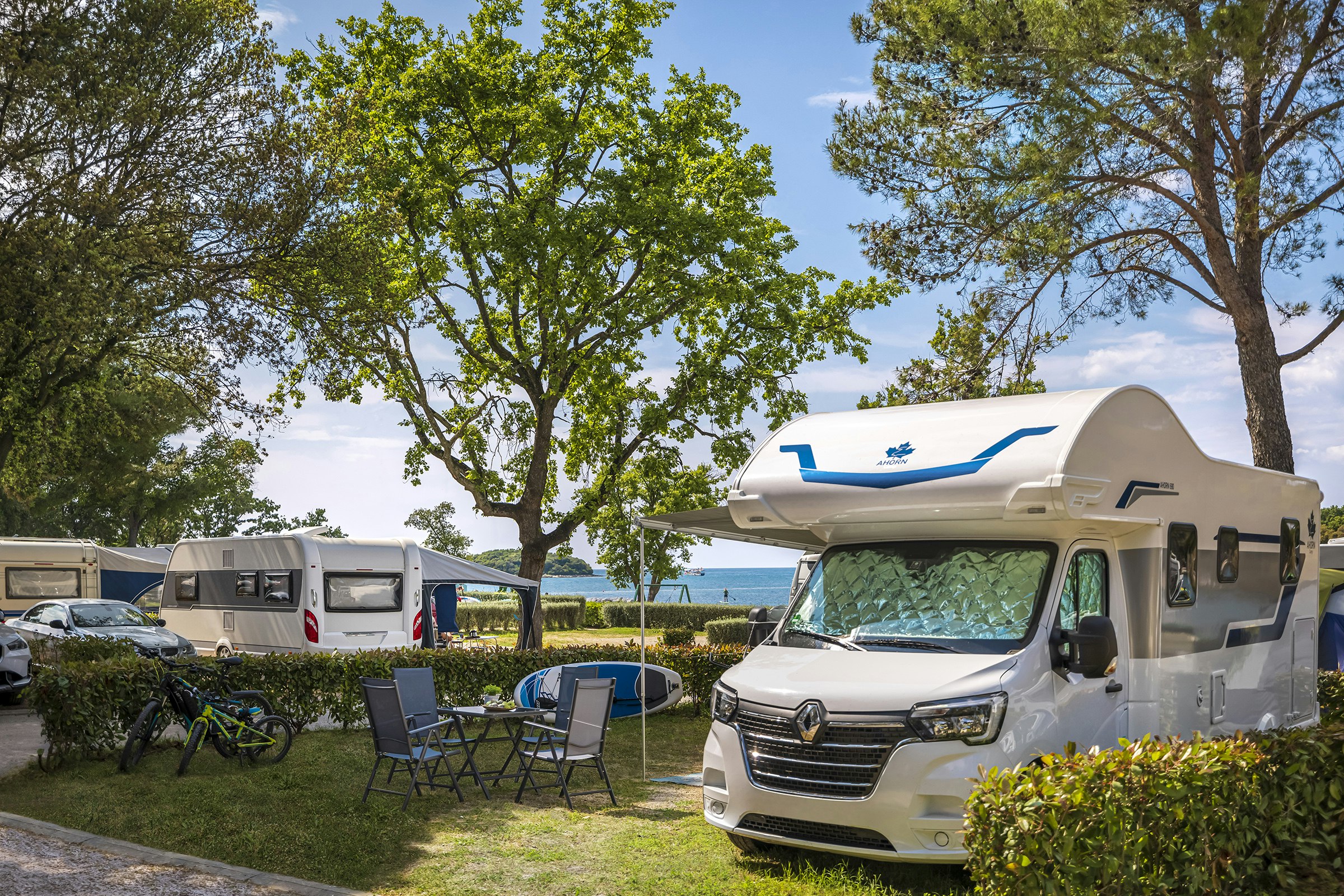 Wohnmobil auf dem Stellplatz mit Markise, Campingstühlen und Fahrrädern im Grünen, daneben weitere Wohnmobile und Bäume.