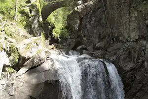 Wasserfall in einer Felsenschlucht mit Bachlauf, umgeben von Bäumen und Felswänden.