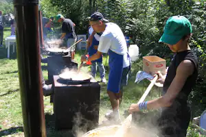 Beim Campinggrillen am Holzkohlegrill: Personen bereiten Essen zu, Rauch steigt auf, im Hintergrund viel Grün und Bäume.