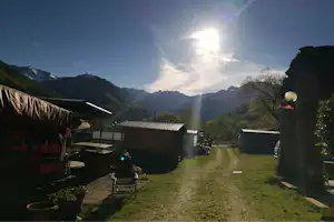 Campingplatz mit Holzunterkünften, Weg und Rasenfläche, im Hintergrund Berge und Sonnenstrahlen durch den Himmel.