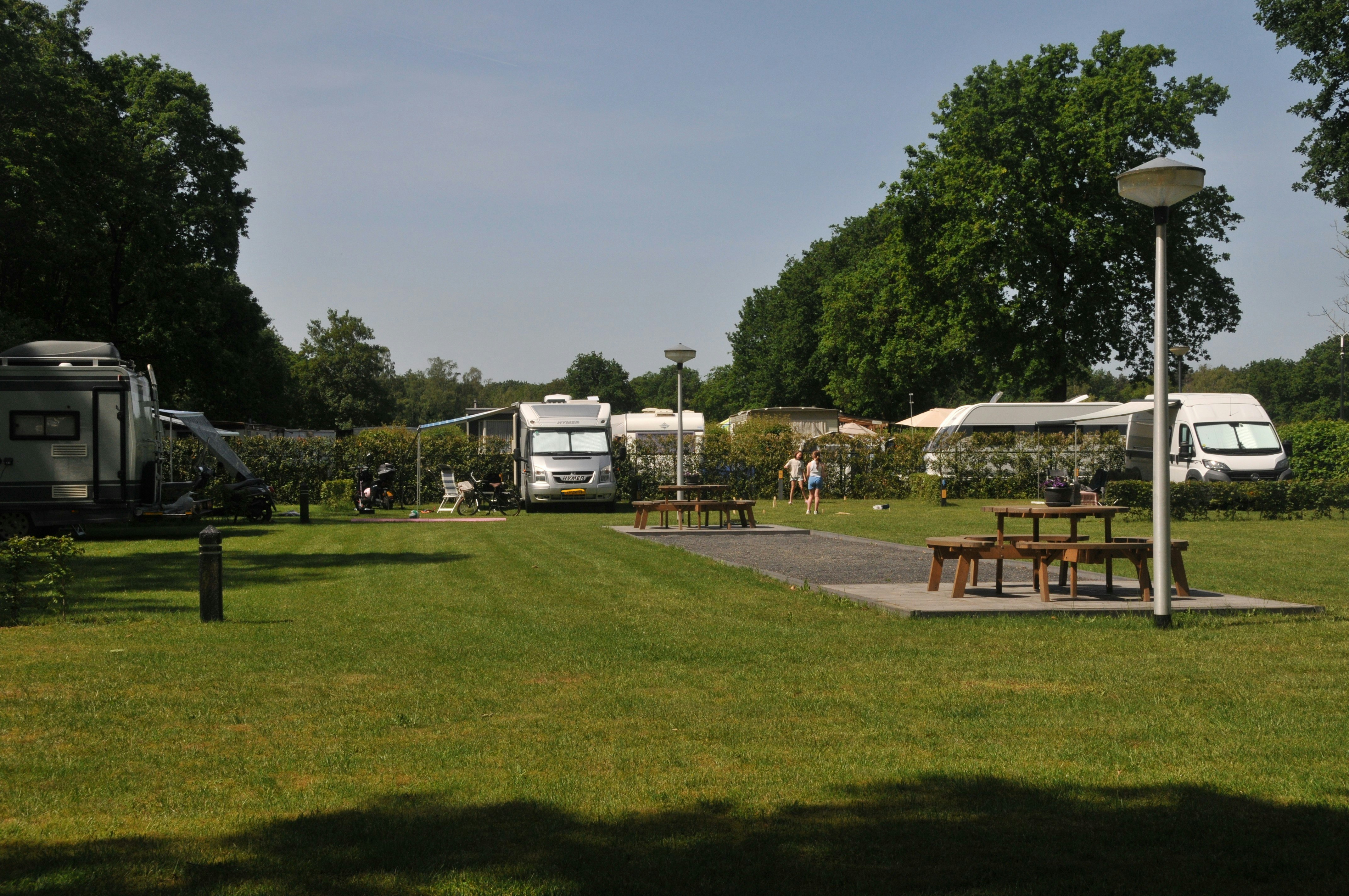 Stellplätze auf einem Campingplatz mit Wohnmobilen und Picknicktischen auf dem Rasen, Bäume und Wege im Hintergrund.