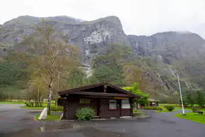 Holzhütte am Campingplatz mit Parkplatz, davor Bäume und Berge im Hintergrund.