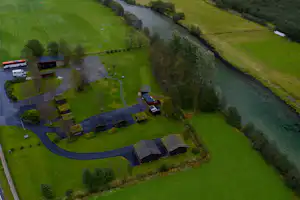 Luftaufnahme eines Campingplatzes mit Stellplätzen, kleinen Gebäuden und Wegen, am Fluss mit grünen Wiesen und Bäumen.