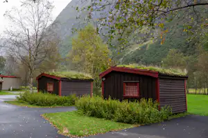 Zwei rote Holz-Blockhütten am Campingplatz, mit gepflastertem Weg, Hecken und Bäumen vor einem grünen Bergtal.