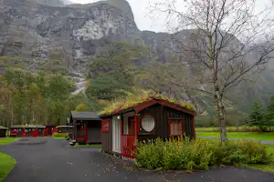 Mobilheim auf einem Campingplatz mit gepflastertem Weg, Hecke und Baum vor hohen Felsbergen im grünen Tal.
