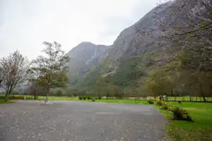 Schotterweg auf einem Campingplatz mit Bäumen und Wiese, im Hintergrund hohe Berge und ein Tal.