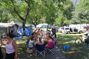 Campingplatz mit Picknicktisch, vielen Personen beim Essen, Zelten und Wohnwagen sowie geparkten Autos im Schatten der Bäume.