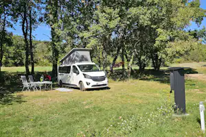 Wohnmobil auf einem Campingplatz im Grünen, daneben Stühle und Tisch unter Bäumen, Schotterweg und Stromsäule.