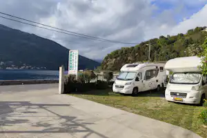 Wohnmobile auf einem Campingplatz an der Einfahrt, Betonweg und Hecke neben einem Schild, Berge im Hintergrund.