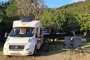 Wohnmobil auf einem Stellplatz mit Markise, zwei Campingstühlen und Tisch, umgeben von Bäumen und Grünfläche.