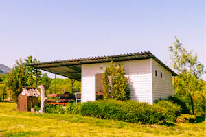 Campinghaus mit Carport, weißer Fassade und grünem Rasen, umgeben von Hecken und Bäumen im Hintergrund.