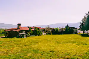 Grüner Rasen vor Campinggebäuden mit roten Dächern, Kamin und Bäumen am Rand, im Hintergrund Berge.