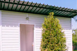 Eingangstür eines Campinghauses an einer weißen Holzfassade, davor Hecke und gepflegter Garten mit Rasen.