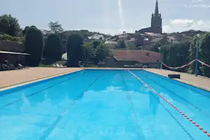Blauer Campingpool mit Bahnen und Blick auf einen Kirchturm über dem Dorf, Bäume und Hecken im Hintergrund.