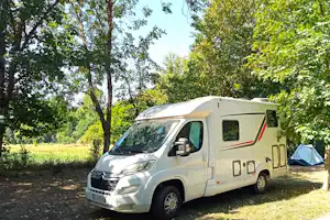 Weißes Wohnmobil auf einem Campingplatz im Grünen, umgeben von Bäumen und mit Zelt im Hintergrund.