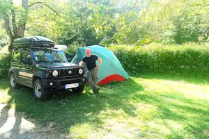 Campingwagen mit Dachbox und Geländewagen neben Zelt auf grünem Stellplatz im Wald, Bäume und Gras im Hintergrund.