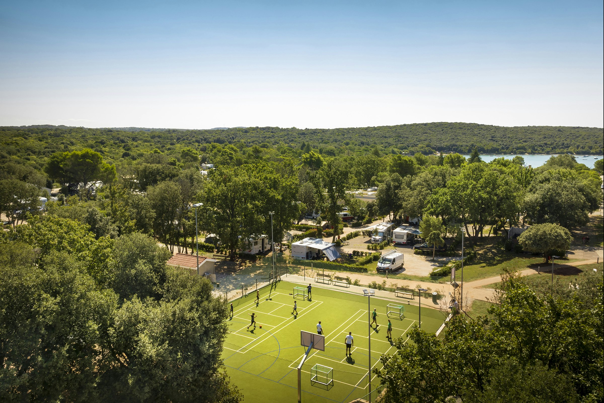 Blick auf eine Campinganlage mit Tennisplatz, Wohnwagen und Autos zwischen Bäumen, im Hintergrund ein See.