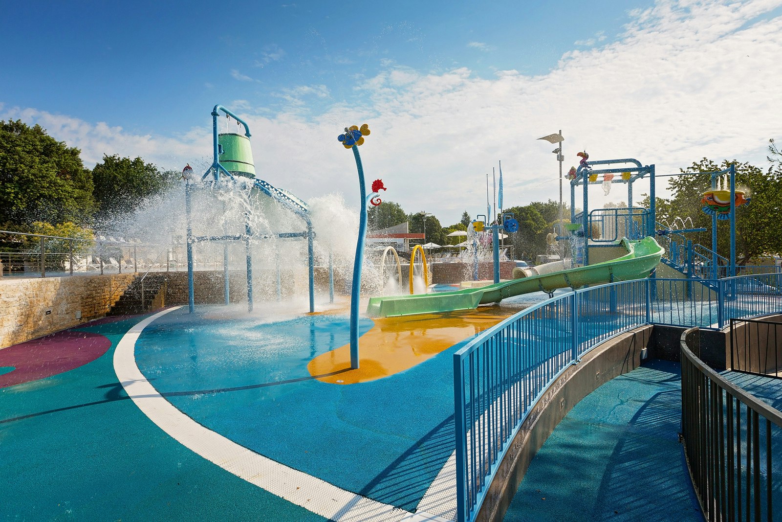 Wasserrutsche und Spritzwasser im Schwimmbecken, blaue Geländer und Spielbereich mit Rutschen auf dem Campingplatz.