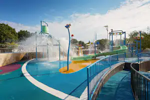 Wasserrutsche und Spritzwasser im Schwimmbecken, blaue Geländer und Spielbereich mit Rutschen auf dem Campingplatz.