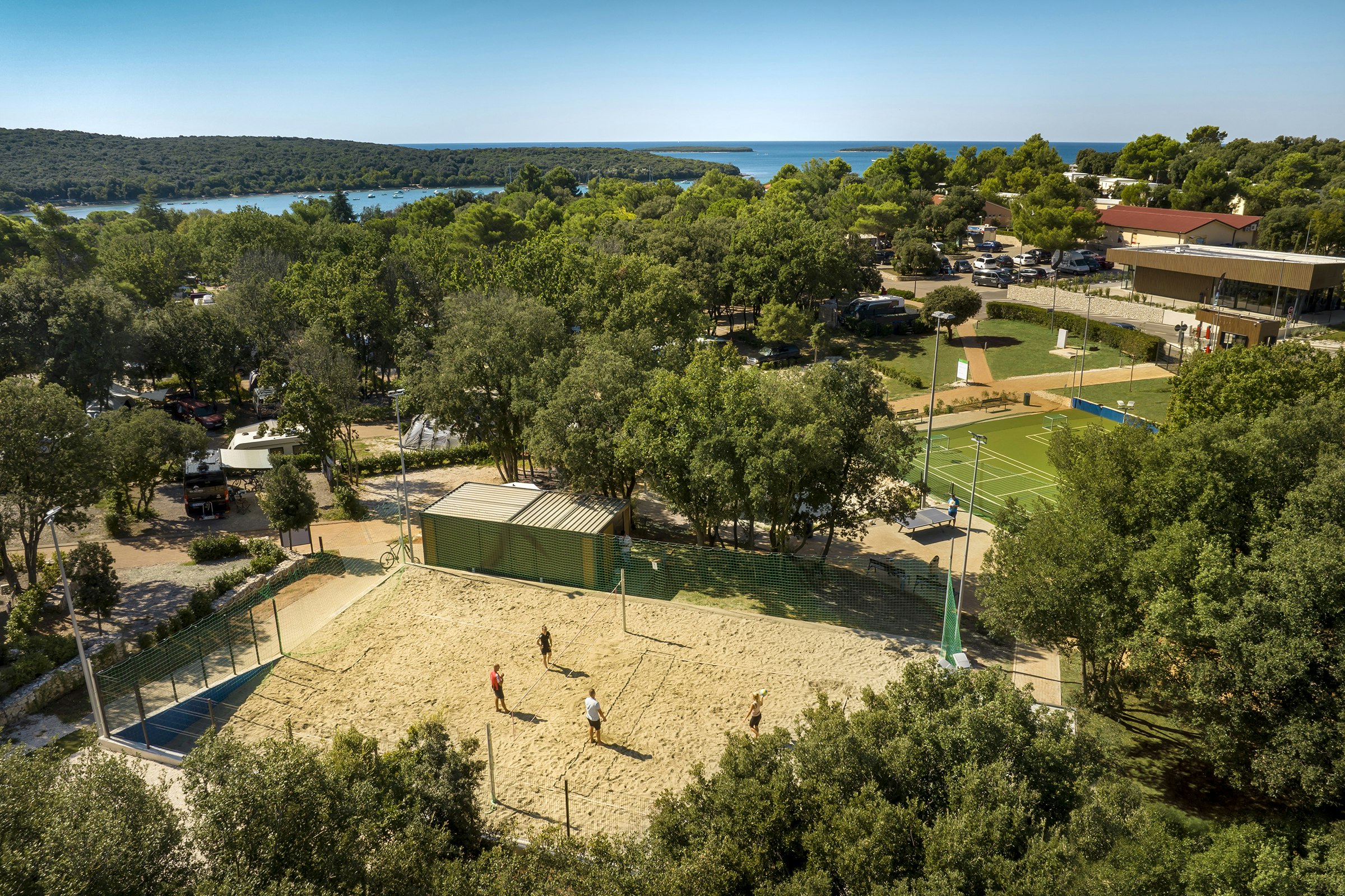 Blick auf Campinganlage mit Sandplatz, Tennisfeld, Bäumen und Parkplatz; im Hintergrund das Meer und eine bewaldete Küste.