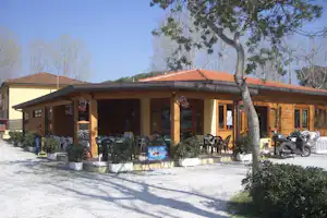 Holzgebäude mit Rezeption und überdachter Terrasse, davor schwarze Gartenstühle und Tische, im Hintergrund Bäume und Rasen.