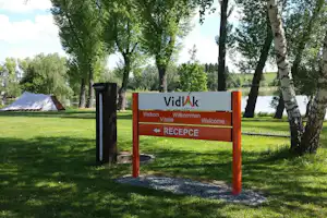 Rezeption-Schild „Vidiak“ auf dem Campingplatz, mit Weg und Rasenfläche zwischen Bäumen und Blick auf den See.