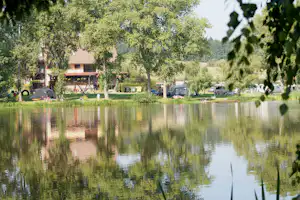 Campingplatz am See: Bungalows mit Terrasse, Zelte und Wohnmobile zwischen Bäumen, im Vordergrund spiegelt sich das Wasser.