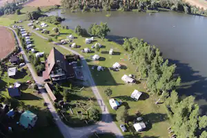 Luftaufnahme eines Campingplatzes am See mit Wohnmobilen, Zelten, Wegen, Bäumen und einem großen Gebäude am Ufer.