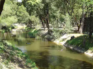 Ruhiger Fluss am Campingplatz mit Uferböschung, Bäumen und Grünflächen, im Hintergrund ein Weg zwischen den Stellplätzen.