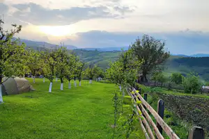 Campingwiese mit Zelt am Rand, Holzzaun, Obstbäume und weite Talsicht bei Sonnenaufgang.