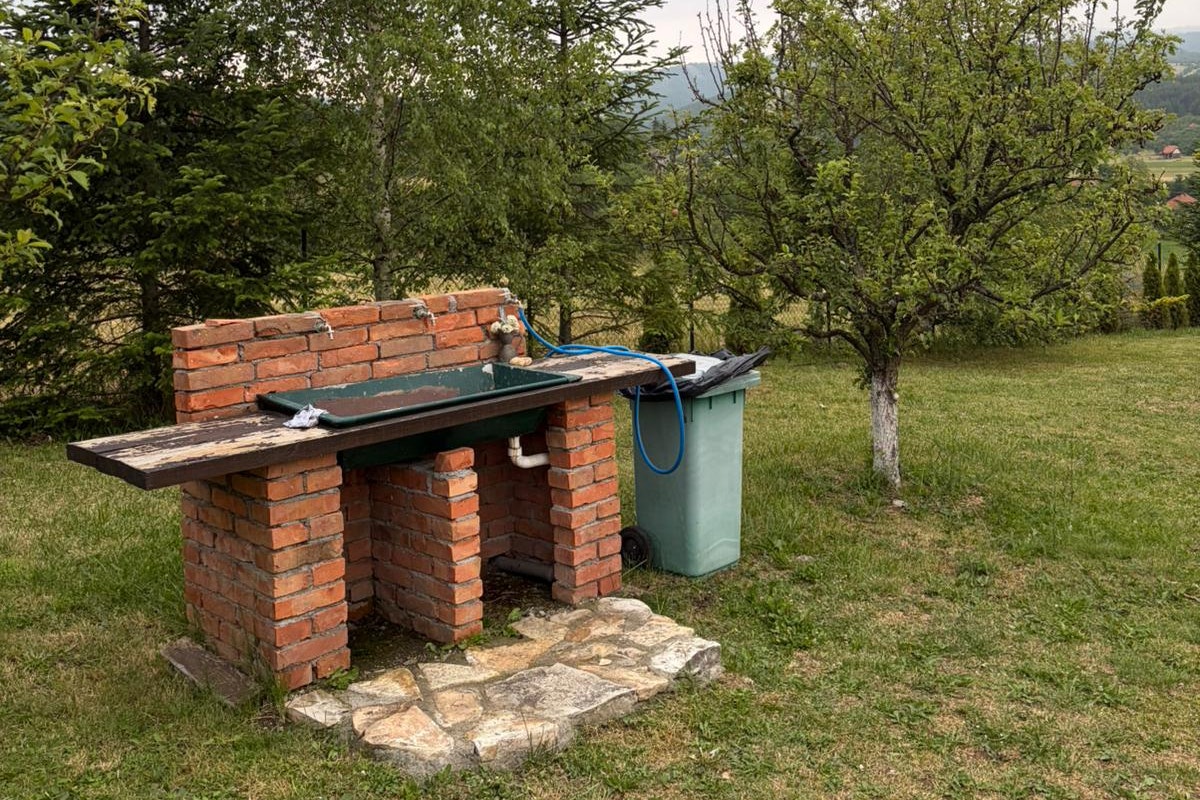 Grillplatz mit gemauerter Ziegelwand, Holzboden und grünem Abfalleimer auf einer Wiese, umgeben von Bäumen.