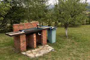 Grillplatz mit gemauerter Ziegelwand, Holzboden und grünem Abfalleimer auf einer Wiese, umgeben von Bäumen.