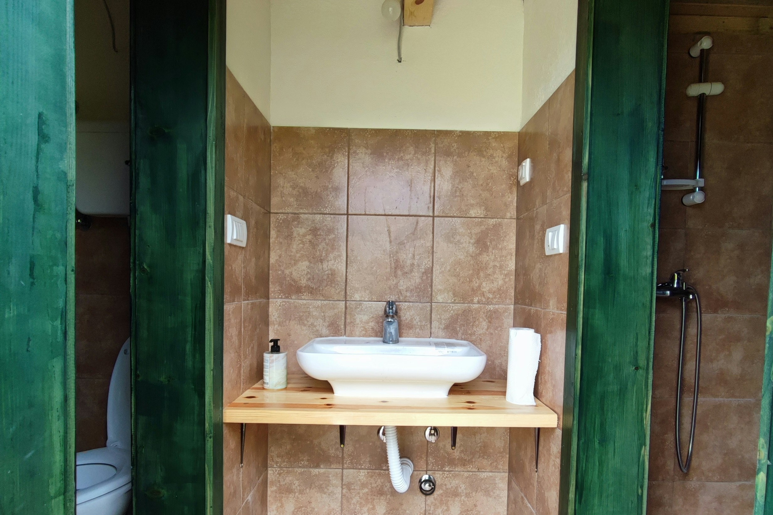 Camping-Badezimmer mit Waschbecken auf Holzplatte, Fliesenboden und grünen Türen, im Hintergrund Toilette und Dusche.
