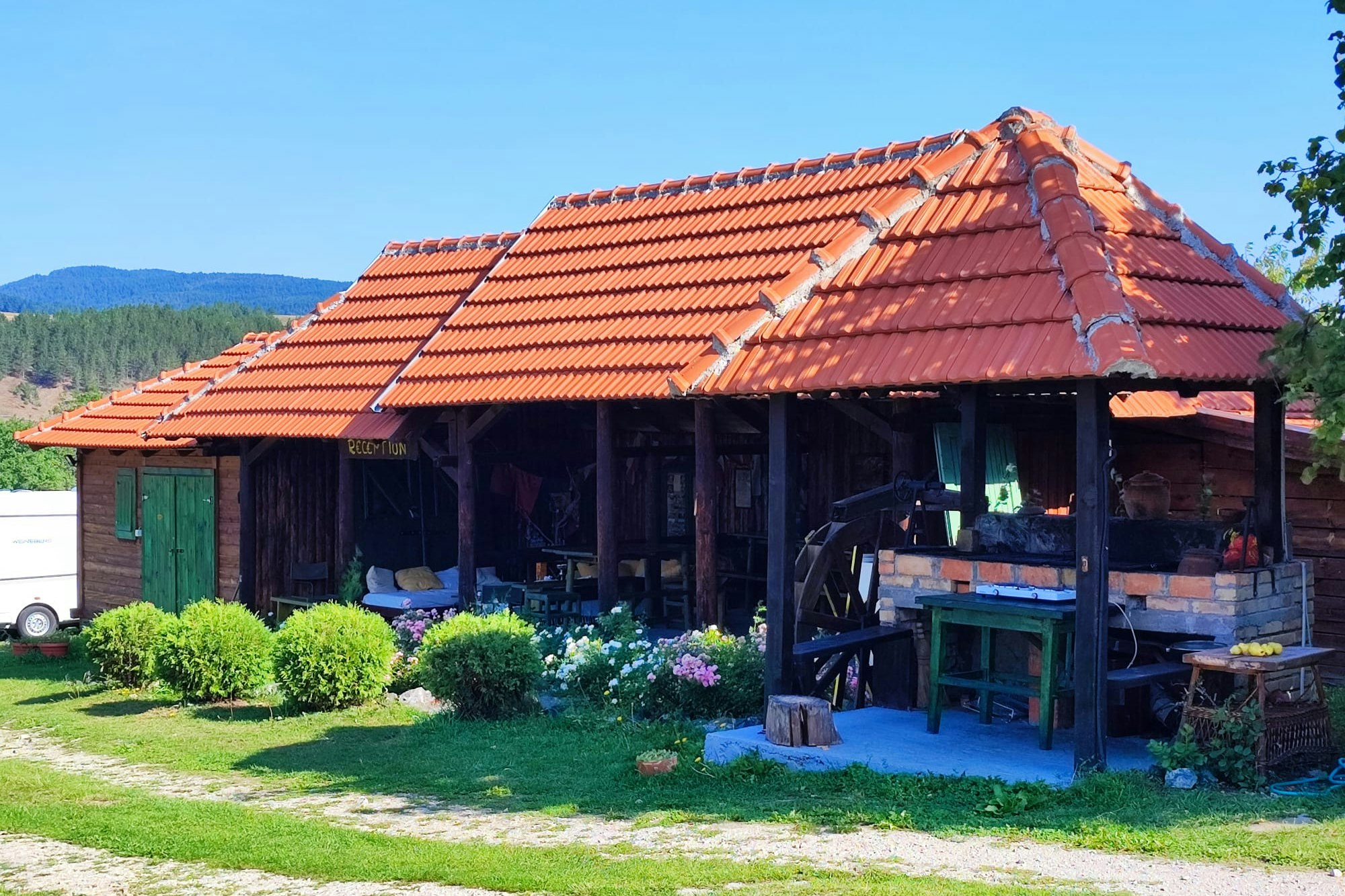 Holzhütte auf dem Campingplatz mit überdachter Terrasse, Holztisch und Bank, gepflegtem Rasen und Büschen im Vordergrund.