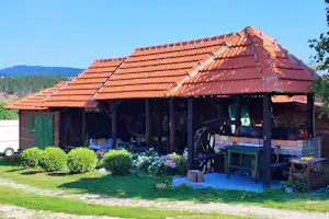 Holzhütte auf dem Campingplatz mit überdachter Terrasse, Holztisch und Bank, gepflegtem Rasen und Büschen im Vordergrund.