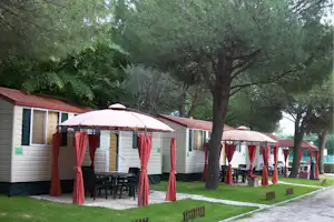 Reihe von Mietzelten auf einem Campingplatz mit roten Vorhängen, Holztischen und Stühlen, umgeben von Bäumen und Rasen.